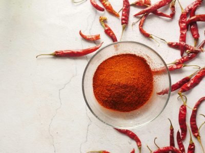 Bowl of vibrant chili powder surrounded by red chili peppers on a textured white background.