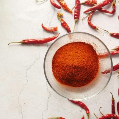 Bowl of vibrant chili powder surrounded by red chili peppers on a textured white background.
